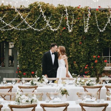 A newly-wed couple kissing infront of a vine covered wall, behind long tables with chairs setup for their wedding reception. Strings of fairy lights are above them.