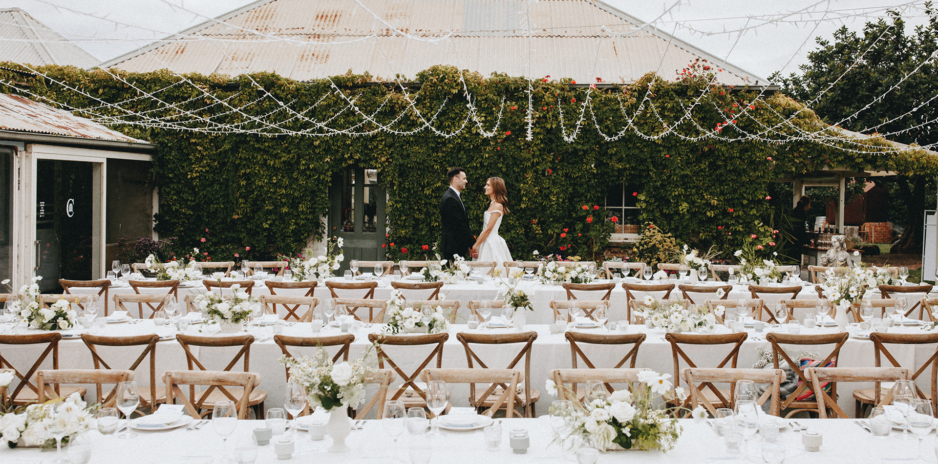 White clothed long tables with timber cross backed chairs, with a bride and groom standing behind them. There are fairy lights overhead, and a vine covered wall in the background.