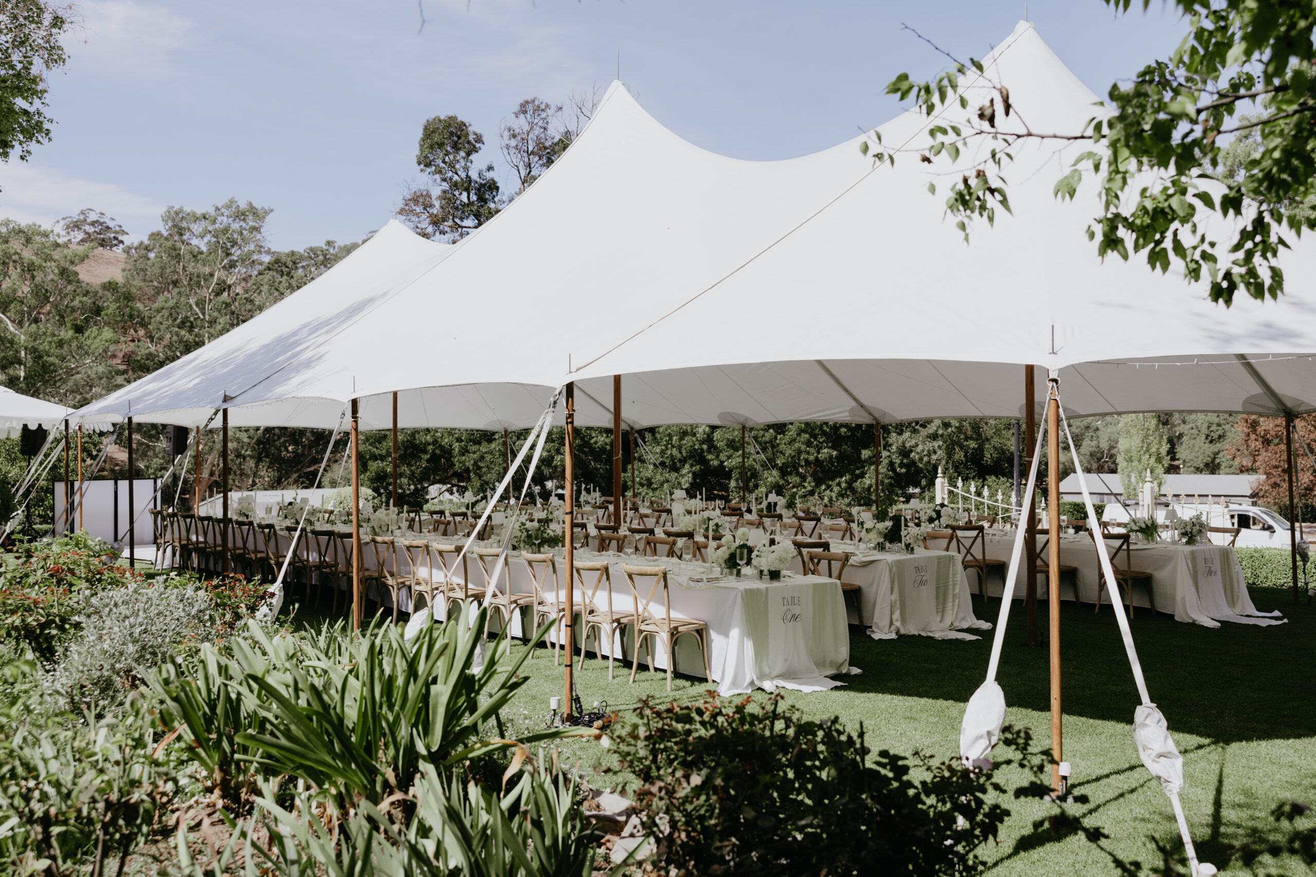 White stretch marquee set up in a garden setting, with long banquet tables and crossback chairs arranged for an elegant outdoor event.
