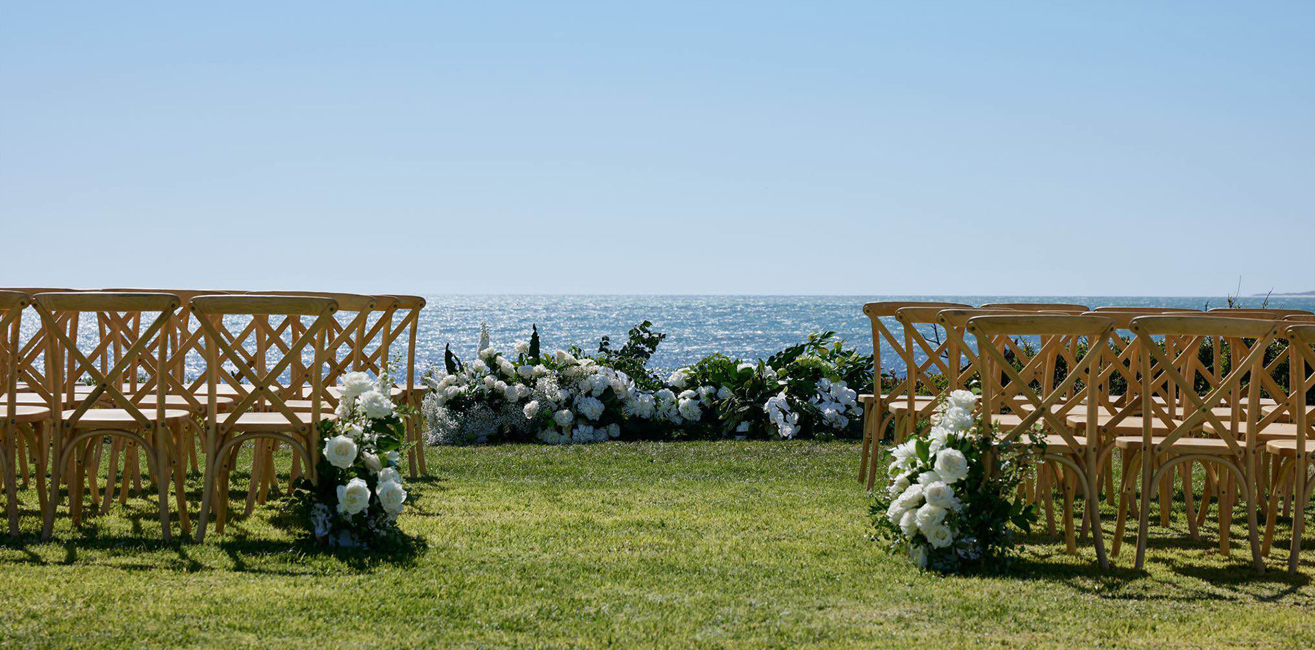 Rows of wooden crossback chairs set up on grass, facing a floral ceremony arrangement with an ocean backdrop under a clear blue sky.