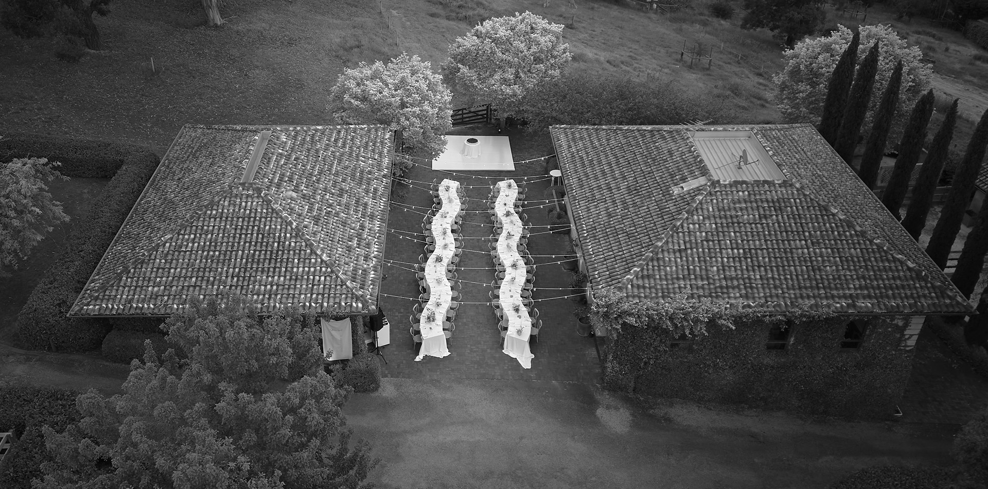 Aerial view of an outdoor event setup between two tiled-roof buildings, featuring long serpentine dining tables with white tablecloths and string lights overhead.