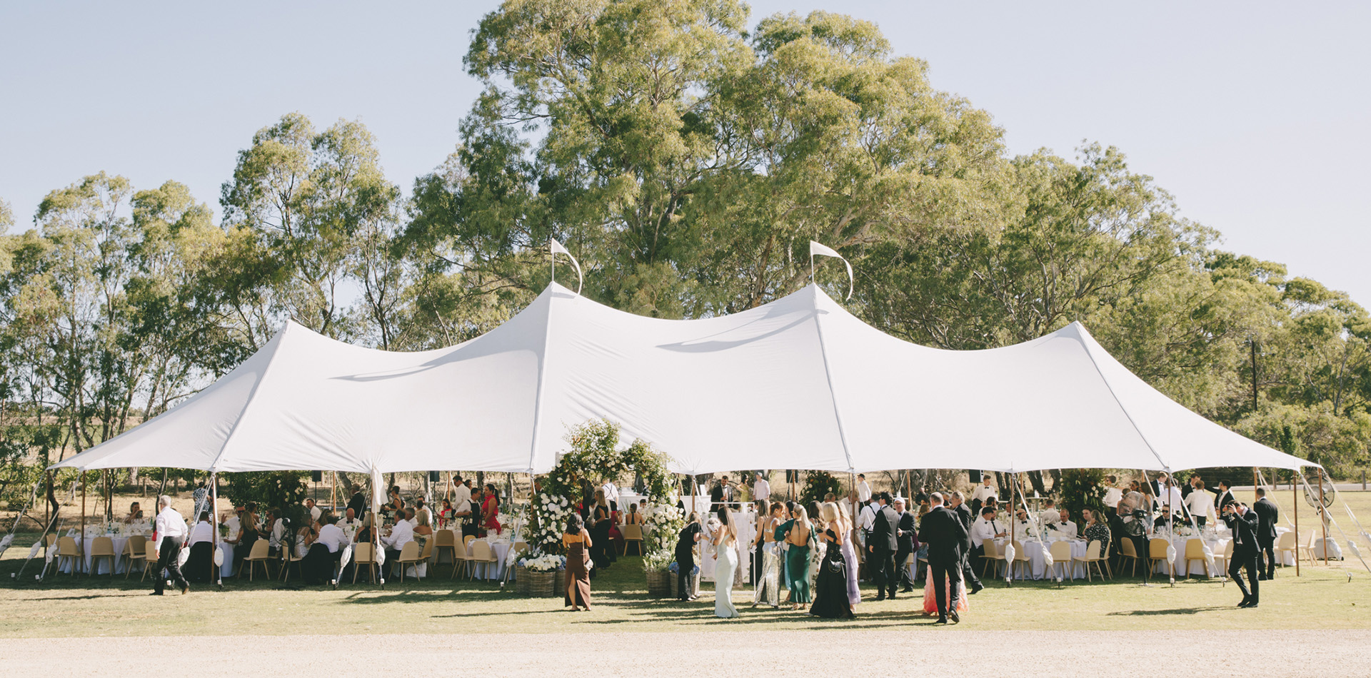 Outdoor event under a large white stretch tent with guests dining and mingling beneath, surrounded by trees and floral decorations